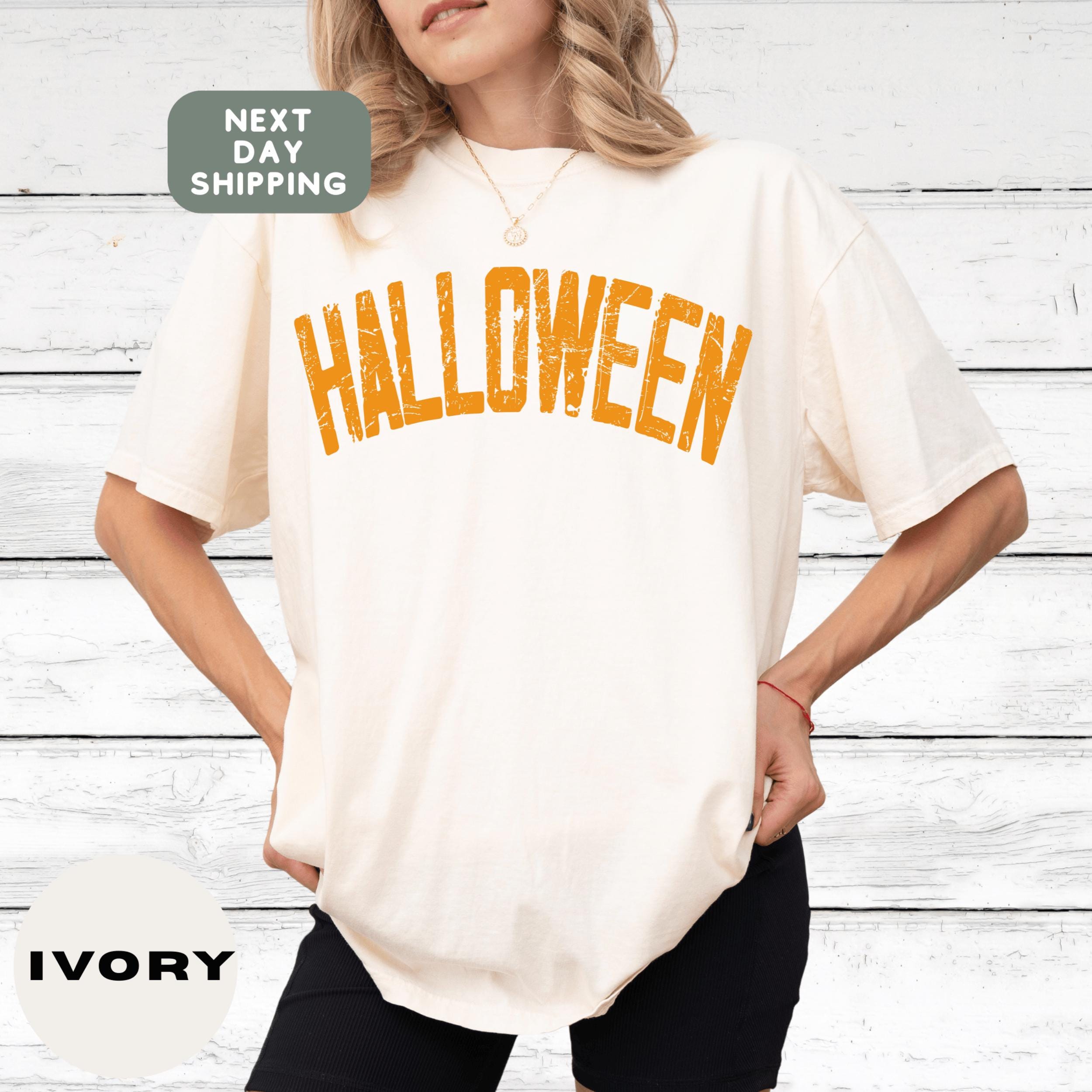 A woman wearing a white t-shirt with the word &quot;Halloween&quot; printed on it, standing in front of a white wooden background.