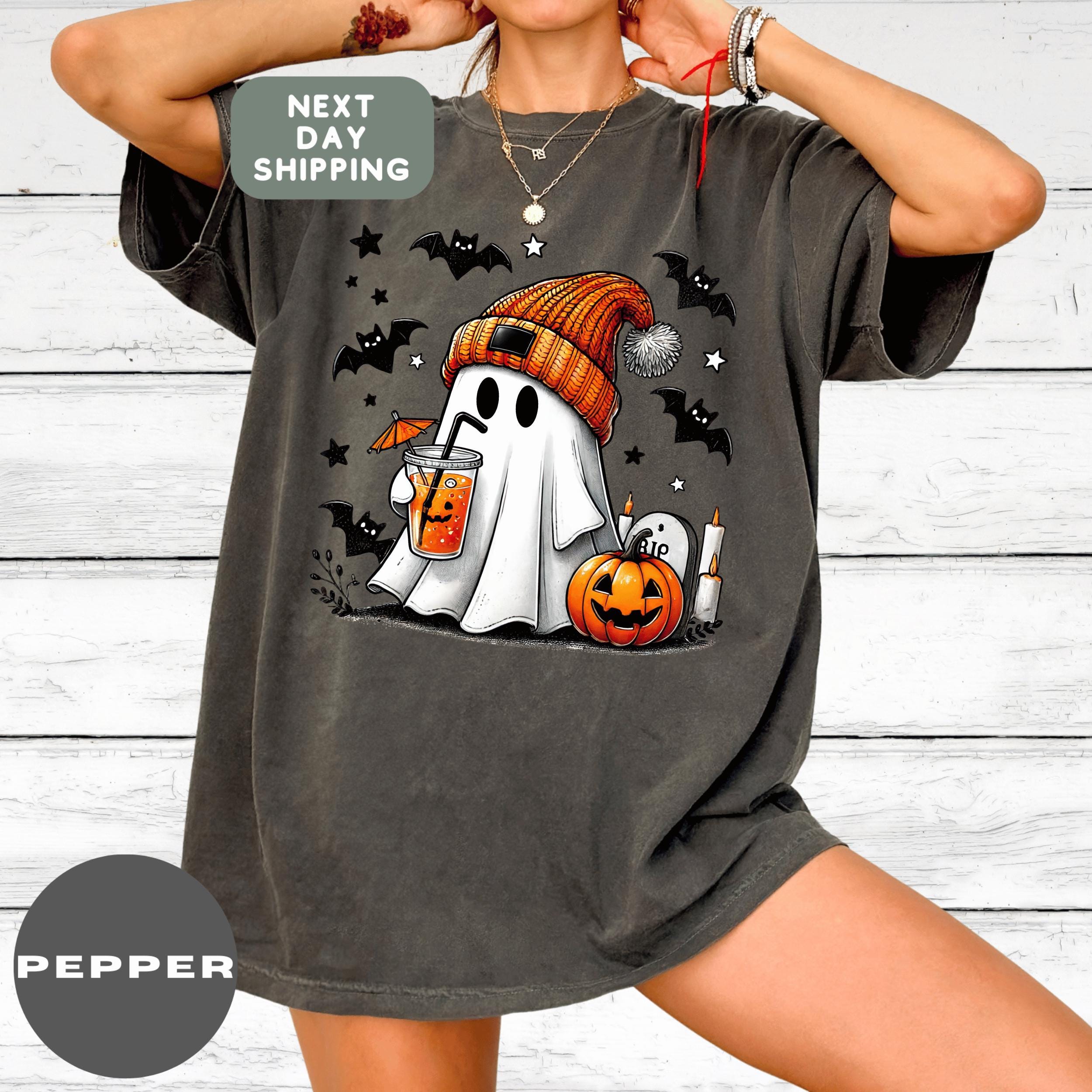 A person wearing a gray t-shirt with a graphic design of a ghost holding a drink and a pumpkin, surrounded by bats and stars.